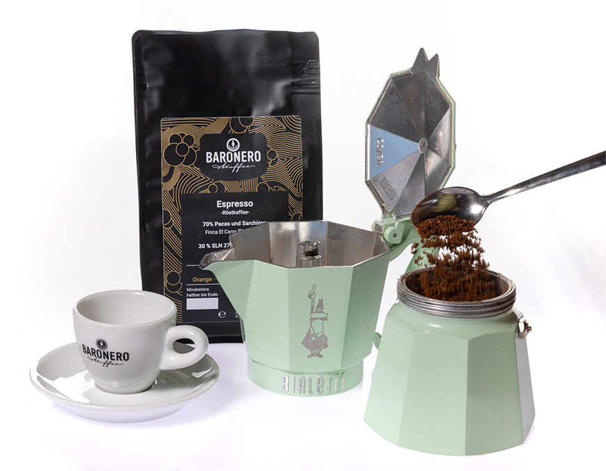 Eine Tüte Baronero Espresso 70-30 mit einer Espressotasse und einer mintgrünen Bialetti-Herdkanne, die gerade mit gemahlenem Kaffee befüllt wird.