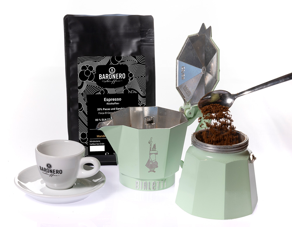 Eine Tüte Baronero Espresso 20-80 mit einer Espressotasse und einer mintgrünen Bialetti-Herdkanne, die gerade mit gemahlenem Kaffee befüllt wird.